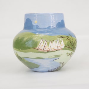 May include: A blue and green ceramic vase with a painted scene of a lake, trees, and a row of white teepees.