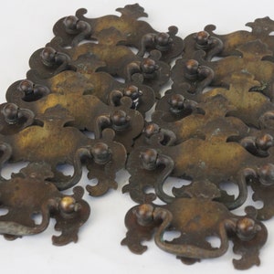 Może przedstawiać: Zestaw 12 antycznych mosiężnych gałek do szafek o ozdobnym, bogatym wzorze. Gałki mają lekko matowe wykończenie i są w dobrym stanie.