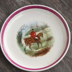 Puede incluir: Un platillo blanco con un borde rosa presenta una ilustraci&oacute;n vintage de una persona montando a caballo a trav&eacute;s de una escena de bosque.
