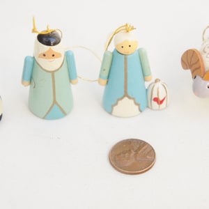 May include: A set of four wooden ornaments including a black and white penguin, two figures in blue and white robes, and a white ram with brown horns, all featuring gold hanging strings, displayed next to a copper-colored United States one-cent coin.