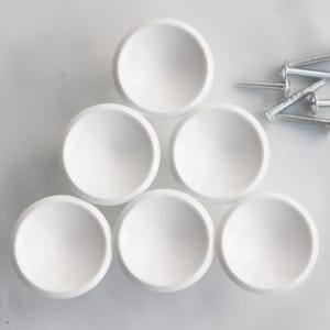 May include: A set of six smooth, matte white circular cabinet knobs arranged in a triangular formation. Each knob features a rounded, dome-like shape. Included with the knobs are six silver-colored metal screws with flat heads for installation.