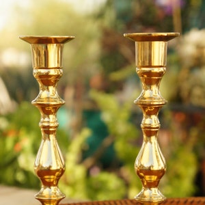 May include: Two gold metal candle holders with a classic design. The candle holders are on a wooden surface.