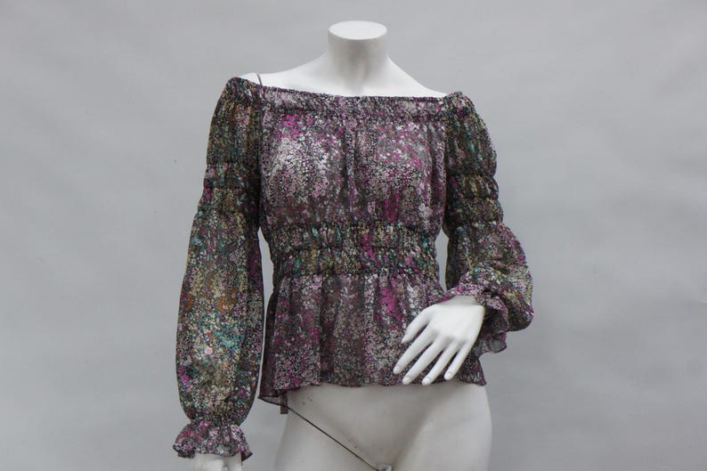May include: Off-the-shoulder blouse with long sleeves. The sheer fabric features a floral pattern in shades of purple, green, and white. The blouse has a gathered waist and elasticated cuffs. The neckline is also elasticated.