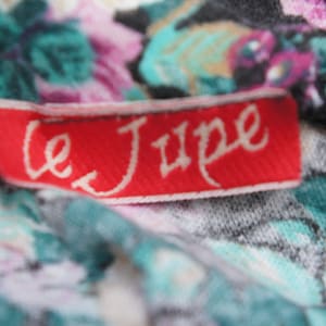 Puede incluir: Una etiqueta de tela roja con texto blanco que dice "Le Jupe" sobre un fondo de tela floral.