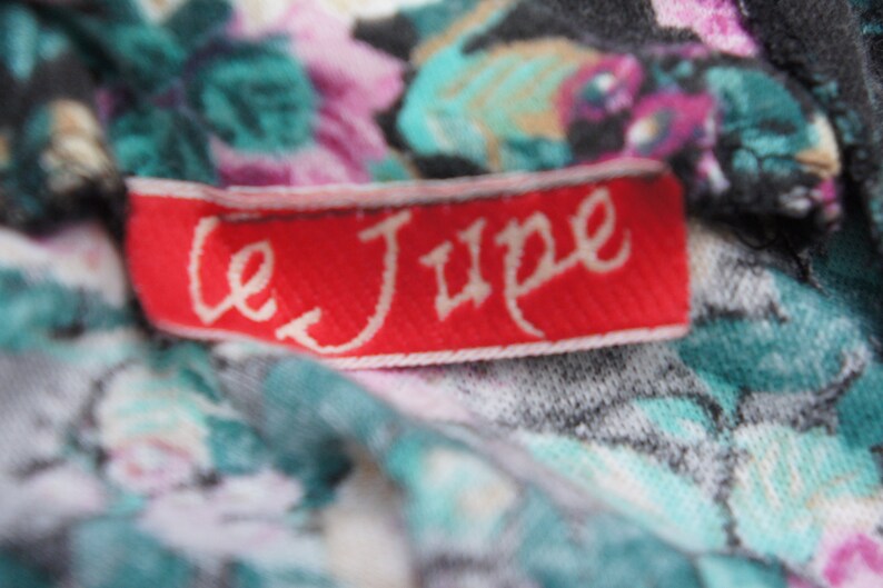 Puede incluir: Una etiqueta de tela roja con texto blanco que dice "Le Jupe" sobre un fondo de tela floral.