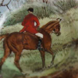 Puede incluir: Una ilustraci&oacute;n vintage de un caballo marr&oacute;n con un jinete que lleva una chaqueta roja y un sombrero negro. El caballo est&aacute; galopando a trav&eacute;s de un campo verde con &aacute;rboles y arbustos en el fondo.