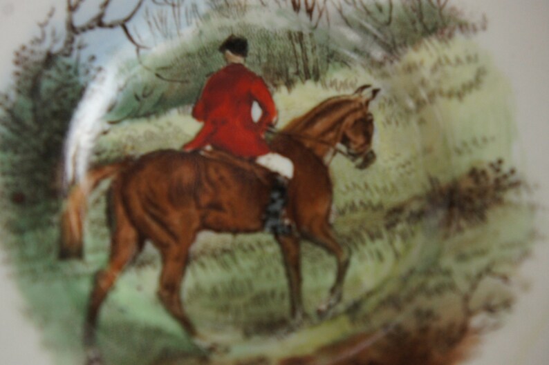 Puede incluir: Una ilustraci&oacute;n vintage de un caballo marr&oacute;n con un jinete que lleva una chaqueta roja y un sombrero negro. El caballo est&aacute; galopando a trav&eacute;s de un campo verde con &aacute;rboles y arbustos en el fondo.