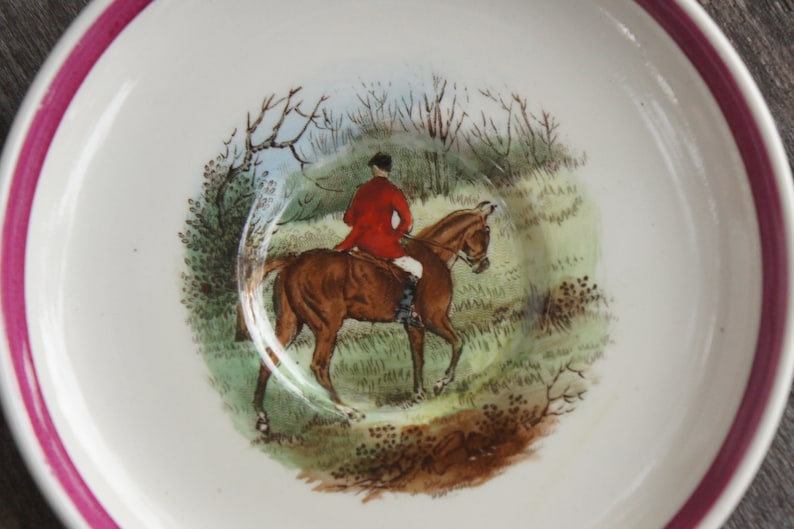 Puede incluir: Un platillo de cer&aacute;mica blanca con un borde rosa presenta una escena pintada a mano de un jinete a caballo en un entorno boscoso. El jinete lleva una chaqueta roja y pantalones de montar negros. El caballo es marr&oacute;n con una estrella blanca en la cara.