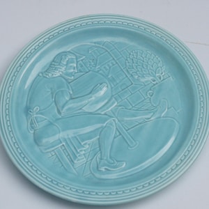 Può includere: Piatto in ceramica azzurra con un rilievo dettagliato di una persona seduta a una scrivania, forse intenta a leggere o scrivere. Il piatto ha un bordo decorativo ed è probabilmente destinato all'esposizione.