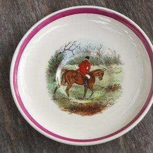 Puede incluir: Un platillo de cer&aacute;mica blanca con un borde rosa presenta una escena pintada a mano de un jinete a caballo en un entorno de bosque. El jinete lleva una chaqueta roja y el caballo es marr&oacute;n.