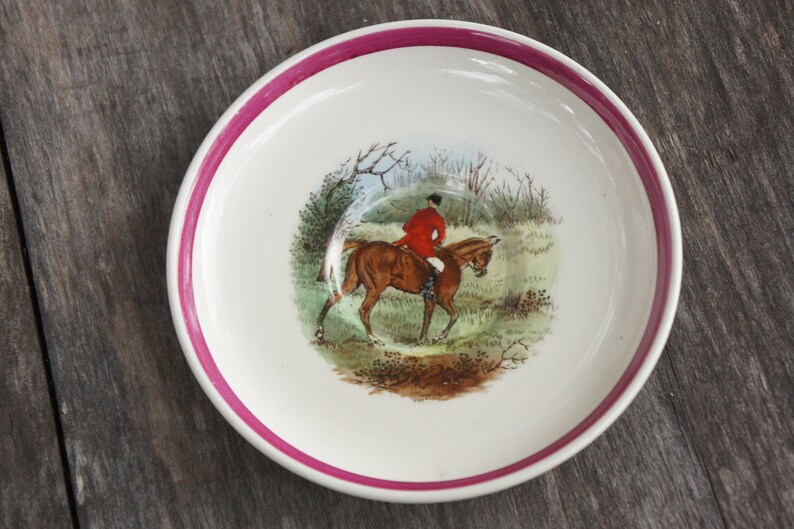 Puede incluir: Un platillo de cer&aacute;mica blanca con un borde rosa presenta una escena pintada a mano de un jinete a caballo en un entorno de bosque. El jinete lleva una chaqueta roja y el caballo es marr&oacute;n.