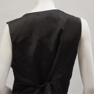 May include: A black vest with a satin finish, viewed from the back. The vest has a V-neck and a tie closure at the waist. The vest is displayed on a mannequin.