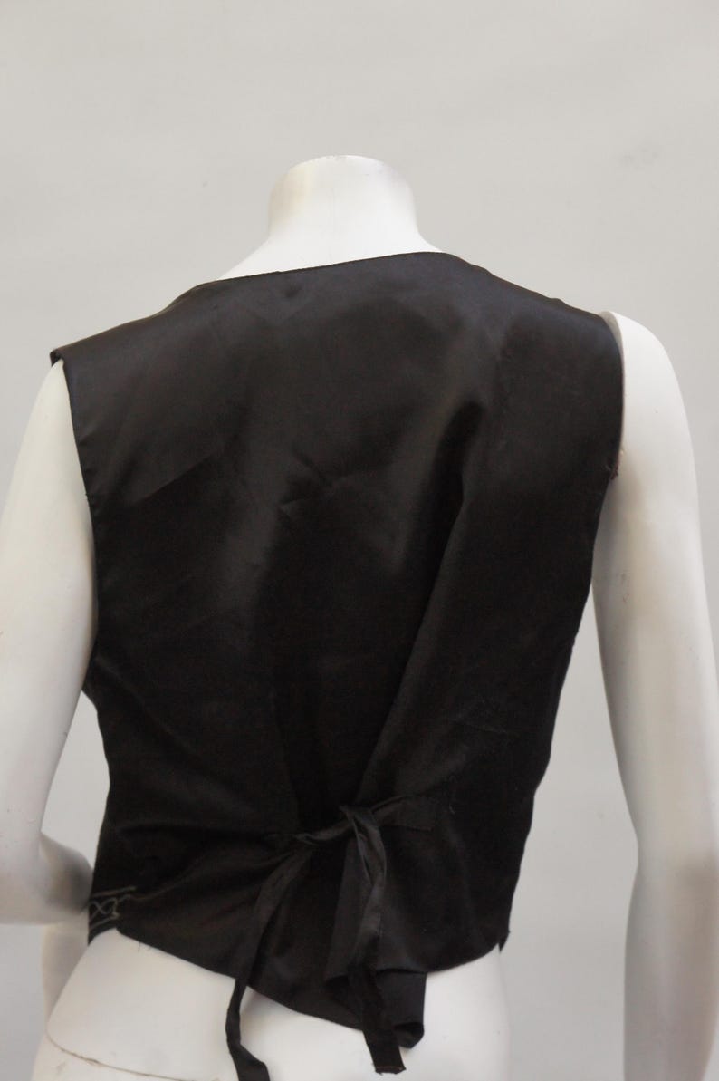 May include: A black vest with a satin finish, viewed from the back. The vest has a V-neck and a tie closure at the waist. The vest is displayed on a mannequin.
