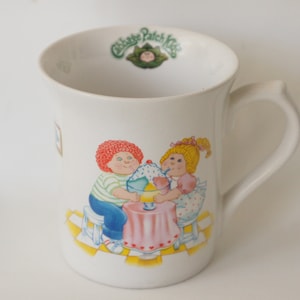 May include: White ceramic mug with a handle. The mug has a colorful illustration of two Cabbage Patch Kids dolls sharing an ice cream sundae. The text "Cabbage Patch Kids" is printed in green on the top of the mug.