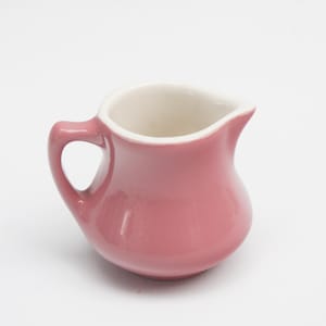 以下が含まれることがあります： 白の内側を持つ、ピンク色の小さなセラミッククリーマー。 湾曲したハンドルと注ぎ口が付いています。 デザインはシンプルでクラシックで、クリームやミルクの提供に適しています。 高さ約10cmです。