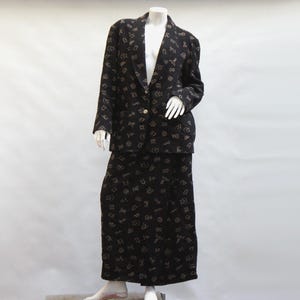 Pode incluir: Um fato de duas peças preto com um blazer e uma saia longa. O fato é coberto por um padrão repetido de símbolos de cor clara. O blazer tem um único botão dourado.