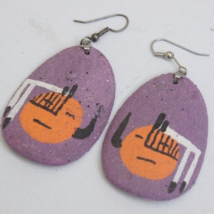Puede incluir: Un par de pendientes morados con un diseño pintado de una cara estilizada con detalles en naranja y blanco. Los pendientes están hechos de arcilla y tienen ganchos plateados.