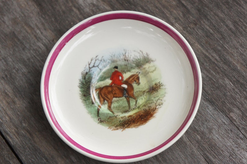 Puede incluir: Un platillo blanco con un borde rosa presenta una ilustraci&oacute;n vintage de una persona montando a caballo a trav&eacute;s de una escena de bosque.
