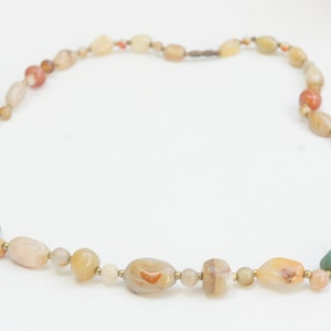 May include: A multi-colored stone necklace with a variety of brown, white, green, and red stones. The necklace is made of natural stones and is strung on a thin gold cord.