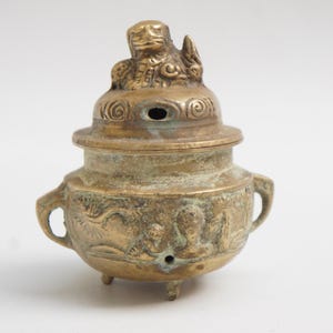 May include: A vintage brass incense burner with a lid featuring a detailed relief of a mythical creature. The burner has a round body with handles and three small feet. The surface shows signs of age and wear.