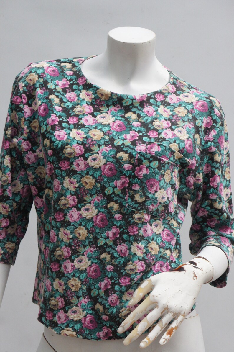 Puede incluir: Una camisa de estampado floral negro y turquesa con un peque&ntilde;o bolsillo en el lado izquierdo. La camisa tiene cuello redondo y mangas tres cuartos.