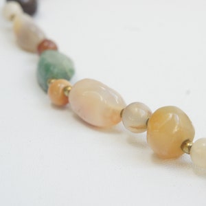 May include: A close-up of a multi-colored gemstone necklace with various shades of brown, green, and orange. The necklace is made of smooth, rounded beads with a gold metal connector between each bead.