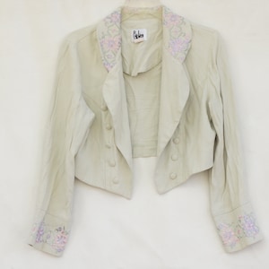 May include: A cropped, light beige jacket with a floral embroidered collar and cuffs. The jacket has a double-breasted closure with buttons and a pointed collar.