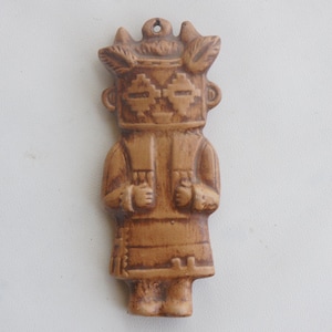 Puede incluir: Figura tallada de color marrón claro de una figura estilizada con tocado, ojos rectangulares y túnica. La figura tiene un agujero en la parte superior para colgar. Los brazos están doblados por los codos, con las manos juntas.