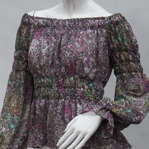 May include: Off-the-shoulder blouse with a floral pattern in shades of purple, pink, and green on a black background. The blouse has long sleeves with ruffled cuffs and a gathered waist. The fabric appears sheer.