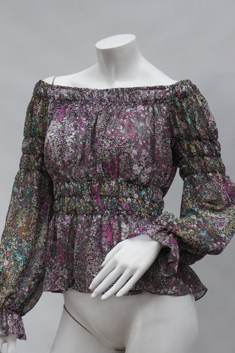 May include: Off-the-shoulder blouse with a floral pattern in shades of purple, pink, and green on a black background. The blouse has long sleeves with ruffled cuffs and a gathered waist. The fabric appears sheer.
