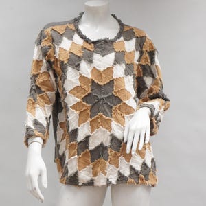 May include: A long-sleeved patchwork top in a starburst pattern. The top features a crew neck and is composed of various fabric patches in shades of gray, white, and tan. The garment is displayed on a mannequin.