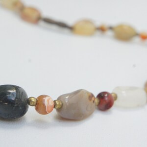 May include: A necklace made of various natural stones, including grey, white, brown, and red. The stones are strung on a brown cord.