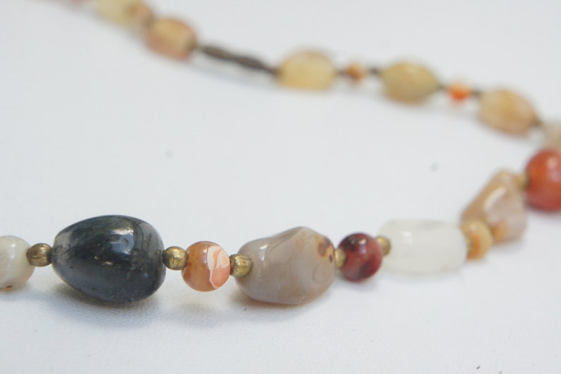 May include: A necklace made of various natural stones, including grey, white, brown, and red. The stones are strung on a brown cord.