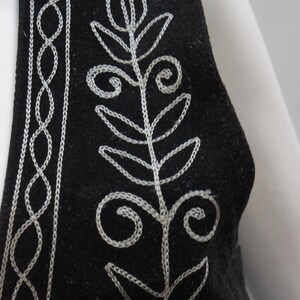 May include: Black vest with white embroidered floral and geometric patterns. The vest has a satin trim along the neckline. The embroidery is detailed and creates a striking contrast against the dark fabric. A stylish piece of clothing.