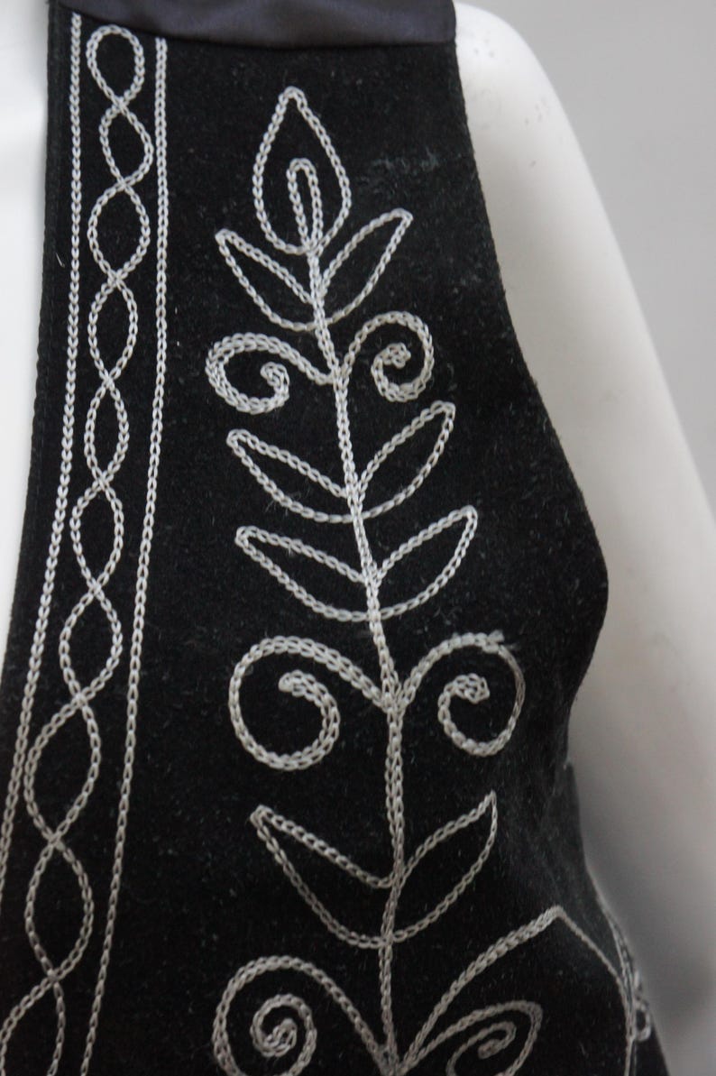 May include: Black vest with white embroidered floral and geometric patterns. The vest has a satin trim along the neckline. The embroidery is detailed and creates a striking contrast against the dark fabric. A stylish piece of clothing.