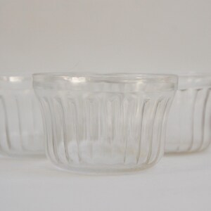 May include: Three clear, ribbed glass containers. The small, round containers have a flared shape and are made of transparent glass. The containers are empty and appear to be vintage or antique.