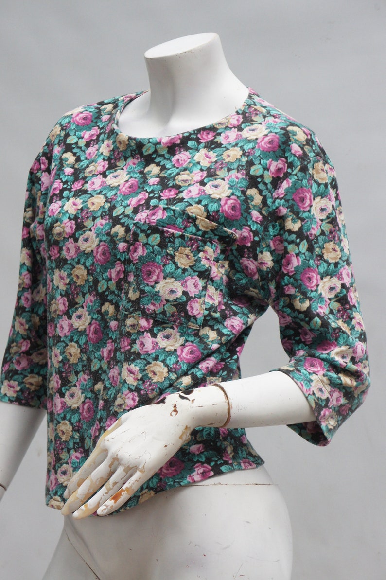 Puede incluir: Una camisa de estampado floral negro con un peque&ntilde;o bolsillo en el lado izquierdo. La camisa tiene cuello redondo y mangas tres cuartos.