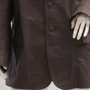 May include: A dark brown leather coat with three buttons and a notched lapel. The coat has a pocket on the right side and a tailored fit. The sleeves are long and the coat appears to be well-worn.