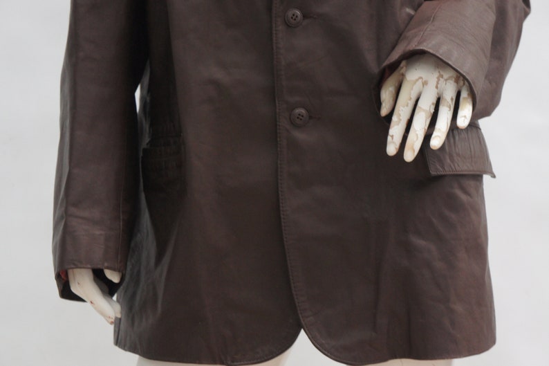 May include: A dark brown leather coat with three buttons and a notched lapel. The coat has a pocket on the right side and a tailored fit. The sleeves are long and the coat appears to be well-worn.