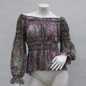 May include: Off-the-shoulder blouse with long sleeves. The sheer fabric features a floral pattern in shades of purple, green, and white. The blouse has a gathered waist and elasticated cuffs. The neckline is also elasticated.