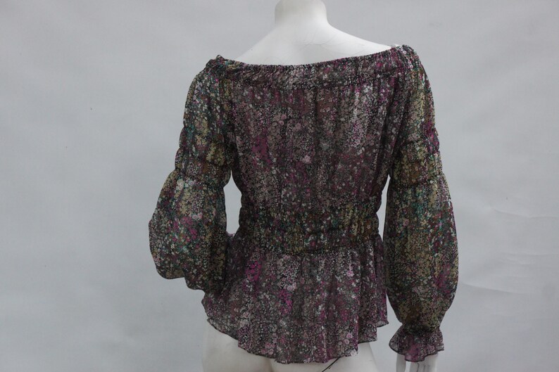 May include: A sheer, off-the-shoulder blouse with long sleeves. The blouse features a floral pattern in shades of purple, green, and yellow. The sleeves are gathered at the wrists, and the waist is cinched with elastic. The blouse has a peplum hem.