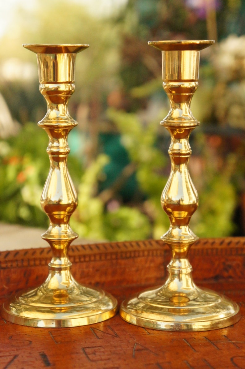 May include: Two gold metal candle holders with a classic design. The candle holders are on a wooden surface.