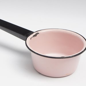 May include: A small, pink enamel saucepan with a black handle. The rim and handle are black, and the interior is a soft pink. The handle has a hole for hanging. The saucepan is likely vintage.