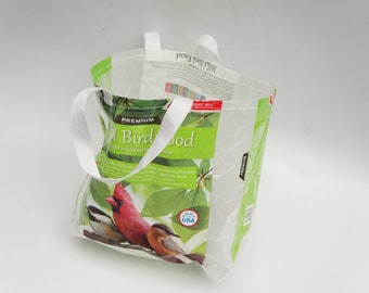 May include: A white reusable bag with white handles. The bag has a green and white label with the text "Premium Bird Food" and a picture of a cardinal, a chickadee, and a downy woodpecker. The label also says "Made in the USA".