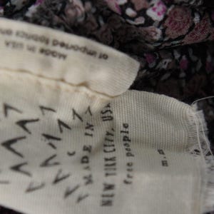 May include: Close-up of a fabric tag with the text "MADE IN NEW YORK CITY, USA" and "free people". The tag is cream-colored with a pattern of black chevrons. The fabric behind the tag has a floral pattern.