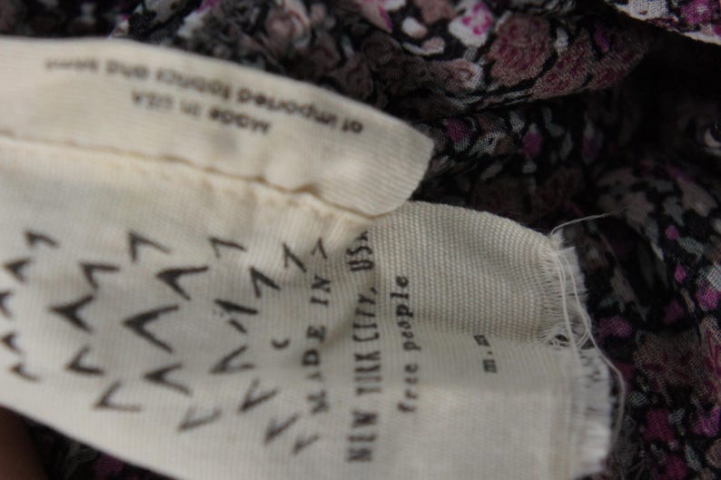 May include: Close-up of a fabric tag with the text "MADE IN NEW YORK CITY, USA" and "free people". The tag is cream-colored with a pattern of black chevrons. The fabric behind the tag has a floral pattern.