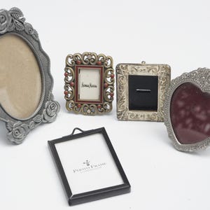 May include: A collection of decorative picture frames in various shapes and styles. Includes an oval frame with rose detailing, a small rectangular frame with the text "Neiman Marcus", and a heart-shaped frame.
