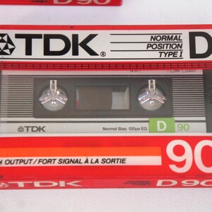 Puede incluir: Una cinta de cassette de audio TDK D90 roja y plateada. La cinta est&aacute; etiquetada como "High Output/Fort Signal &agrave; la Sortie" y "Normal Bias 120&micro;s EQ".