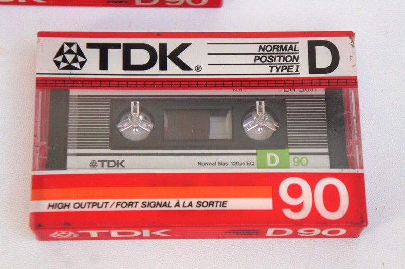 Puede incluir: Una cinta de cassette de audio TDK D90 roja y plateada. La cinta est&aacute; etiquetada como "High Output/Fort Signal &agrave; la Sortie" y "Normal Bias 120&micro;s EQ".