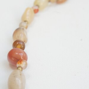 May include: A close-up of a necklace made of natural stone beads. The beads are various shades of brown, orange, and white. The necklace is strung on a thin cord.